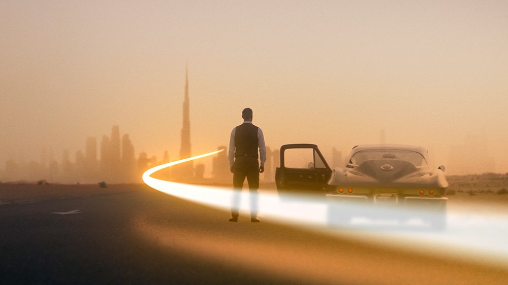 A person stands on a highway at sunset with a vintage sports car parked nearby, city skyline in the background, and a trail of light leading into the distance.