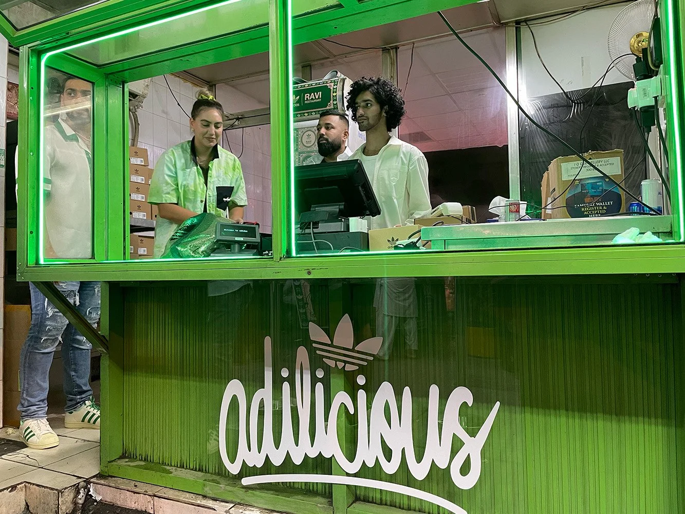A green food stall with the word 'addicious' and a stylized leaf logo, where three people are working and a woman is ordering, inside a restaurant or food court.