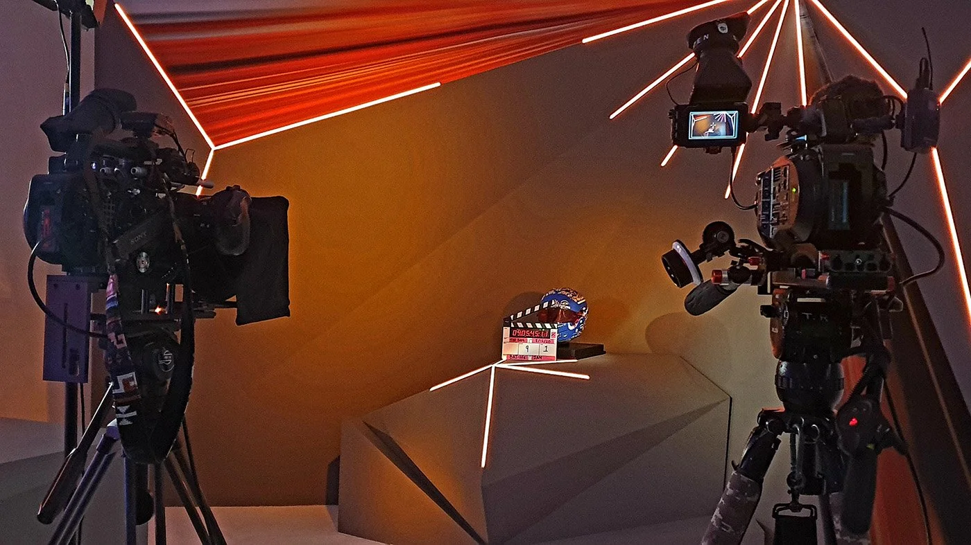 Two professional cameras on tripods are set up to film a scene with a futuristic yellow and orange geometric backdrop, featuring creative lighting and a small shelf with a fantasy-themed globe, dice, and a clapboard.