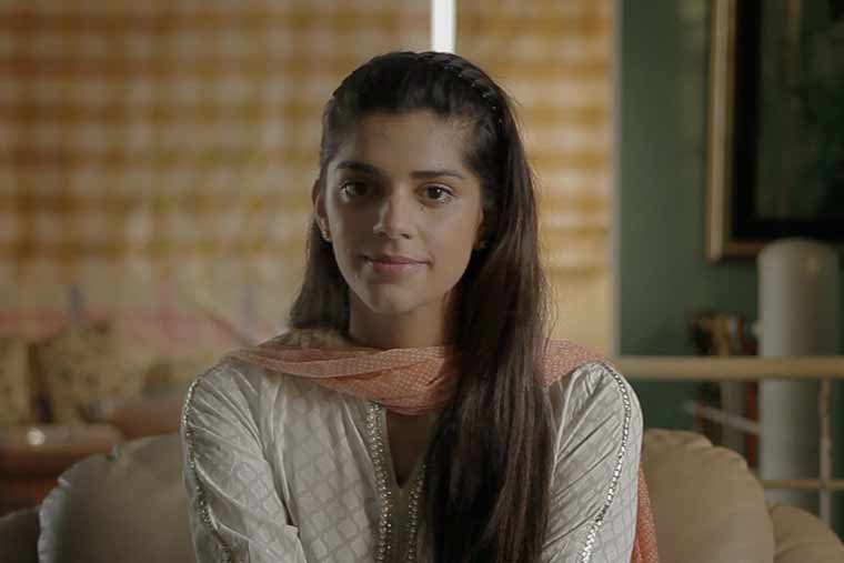 Young woman with long dark hair sitting on a beige sofa in a room with wooden blinds and artwork hanging on the wall.