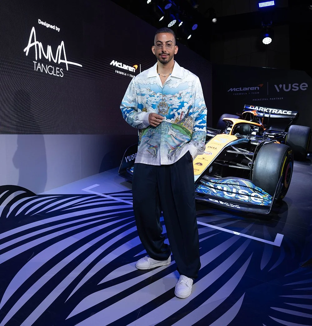 A man standing next to a yellow Formula 1 race car at an event, with a large screen behind him displaying the text "Designed by Anna Tangles" and the McLaren logo.