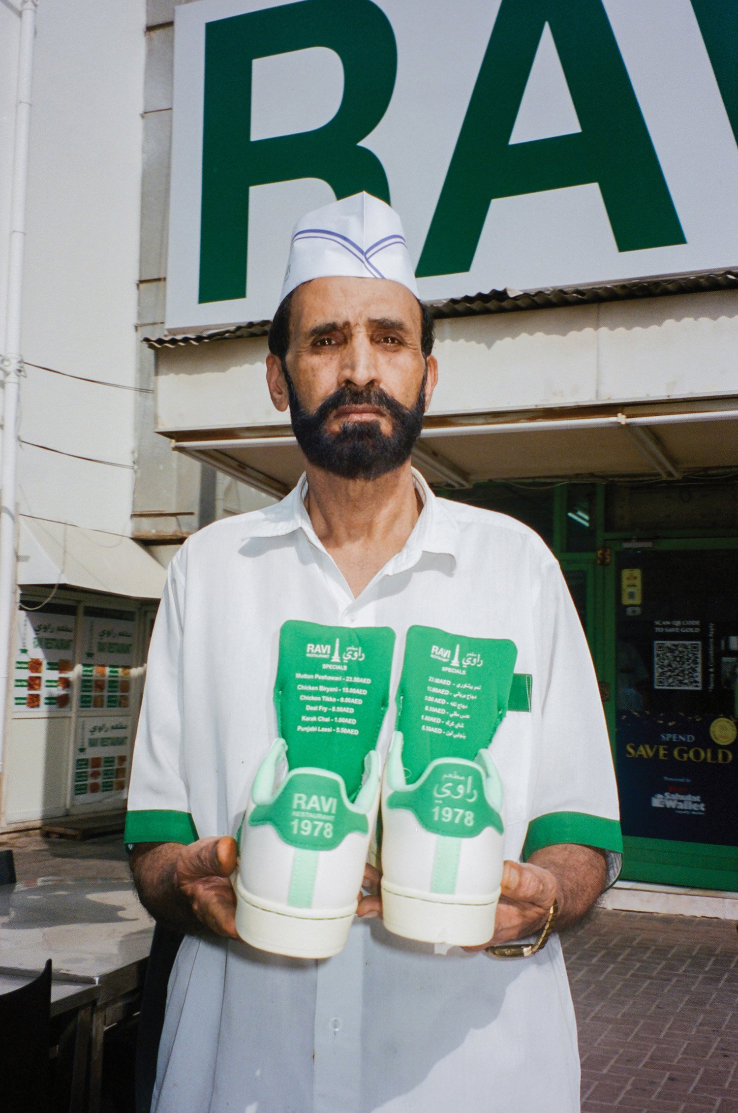 A man with a beard wearing a white shirt and white cap standing outside a store, holding two foam gloves with a green logo and a list of menu items, in front of a large green and white sign.