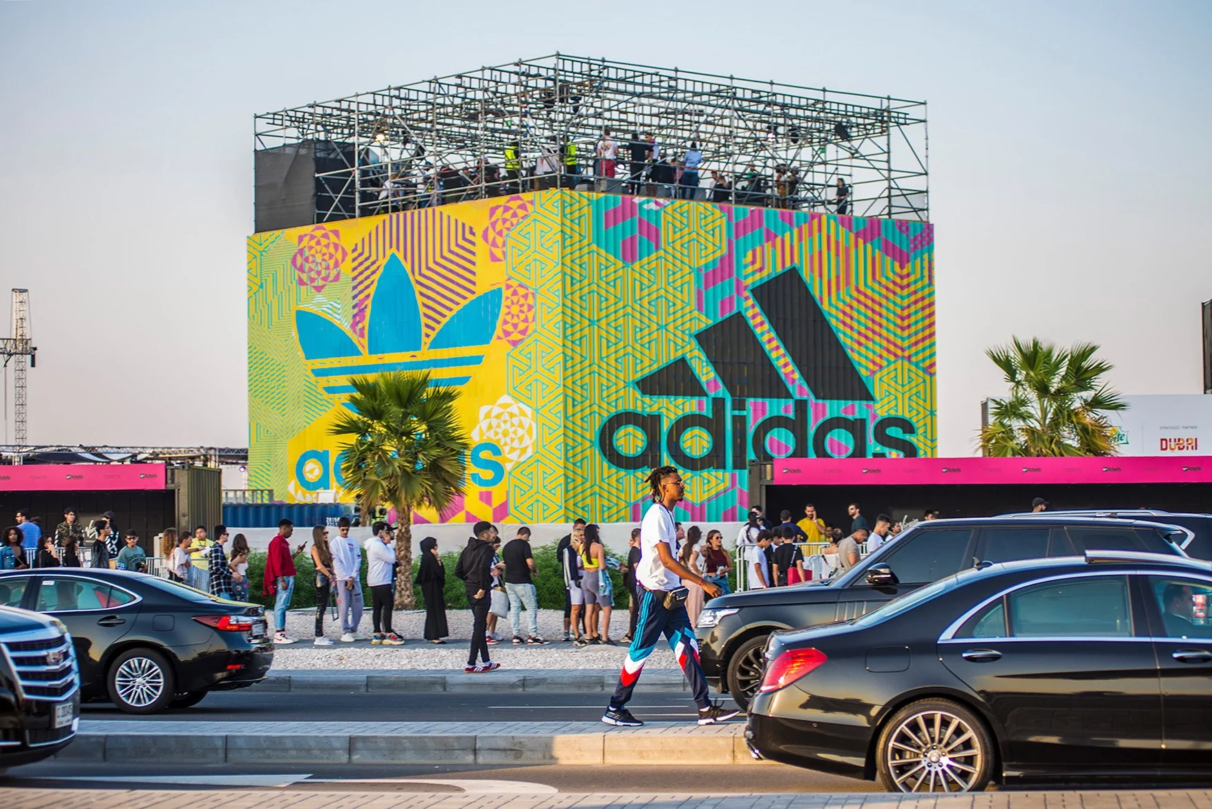 A busy city street with multiple people walking past a large colorful Adidas billboard featuring a vibrant and intricate design.