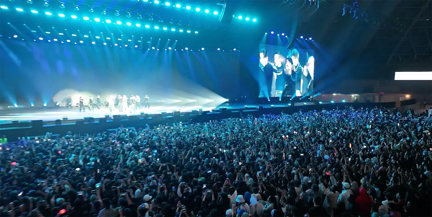 Large indoor concert arena filled with a crowd of people watching a stage performance with bright blue lights and a giant screen displaying the performers.