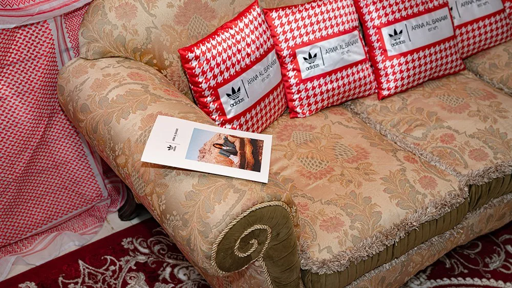A sofa with ornate floral upholstery and fringe along the edges, adorned with three red and white houndstooth patterned Adidas bags and a brochure on the armrest.