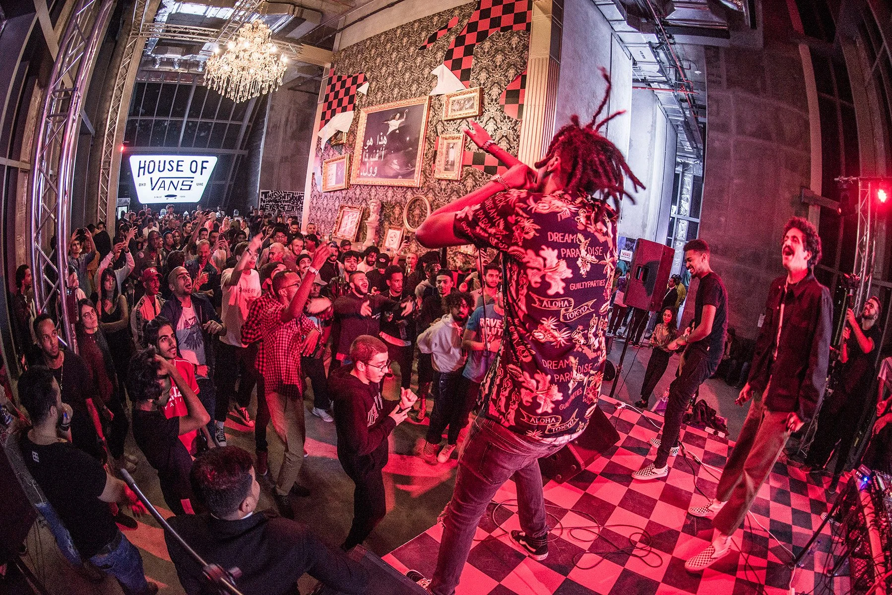 A performer with dreadlocks and a colorful shirt is on stage pointing towards the audience, who are dancing and watching attentively at an indoor concert or event. The venue has ornate wall decorations, framed pictures, and a large chandelier, with a sign that reads 'House of Vans' in the background.