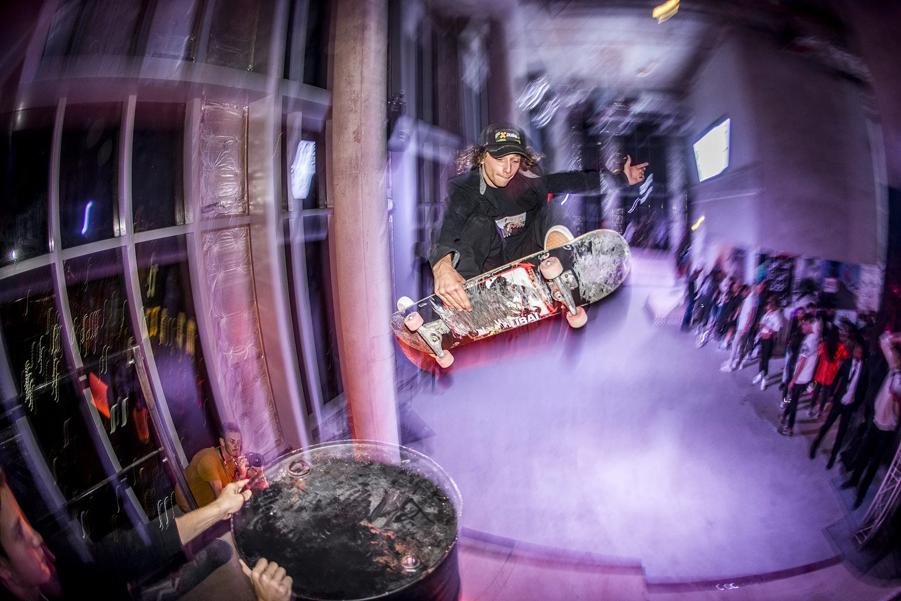 A skateboarder is performing an air trick, jumping off a ledge indoors with a glass of drink in the foreground. Spectators are watching, and the environment is lively with colorful lighting and motion blur.