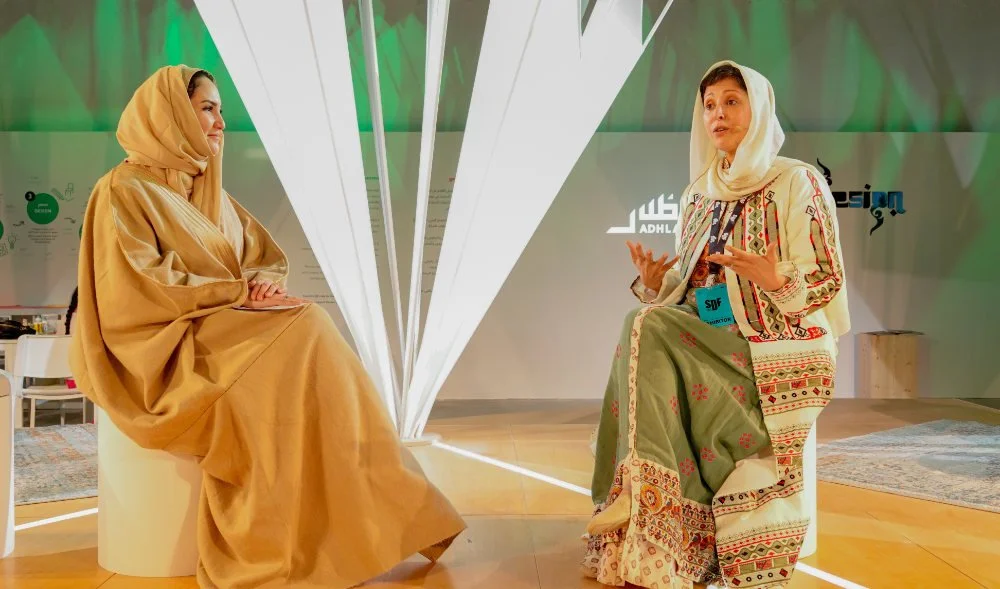 Two women wearing traditional clothing and headscarves are engaged in conversation indoors. One woman is seated on a white stool, while the other is speaking and gesturing with her hands.