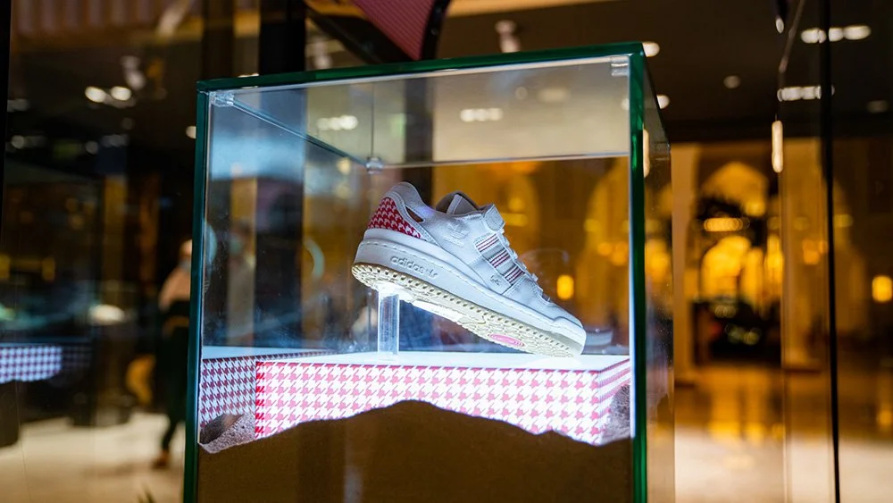 A white Adidas sneaker displayed inside a glass case with a patterned fabric base at a store or exhibition.