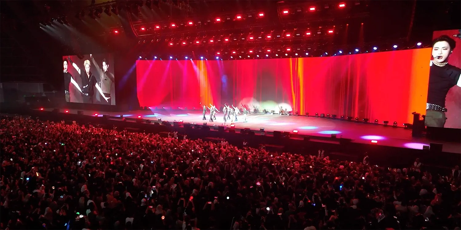 A large concert stage with bright red, orange, and purple lighting, featuring a group of performers dancing and singing. A big screen images three performers on the left and a close-up of one performer on the right, with an audience watching and recording with their phones.