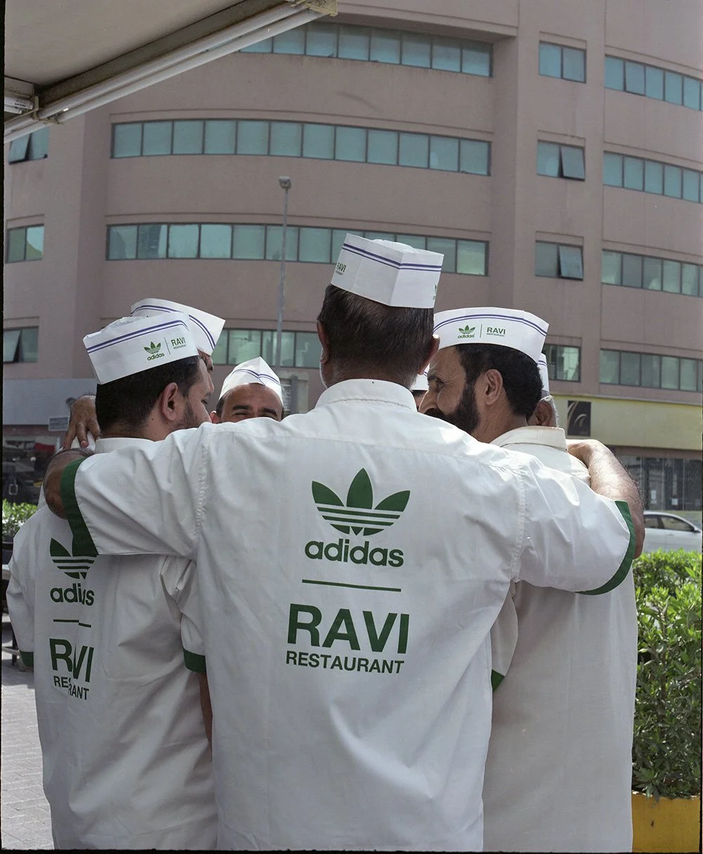 Group of restaurant workers wearing white uniforms with Adidas and RAVI Restaurant logos, standing in a huddle outside a building.