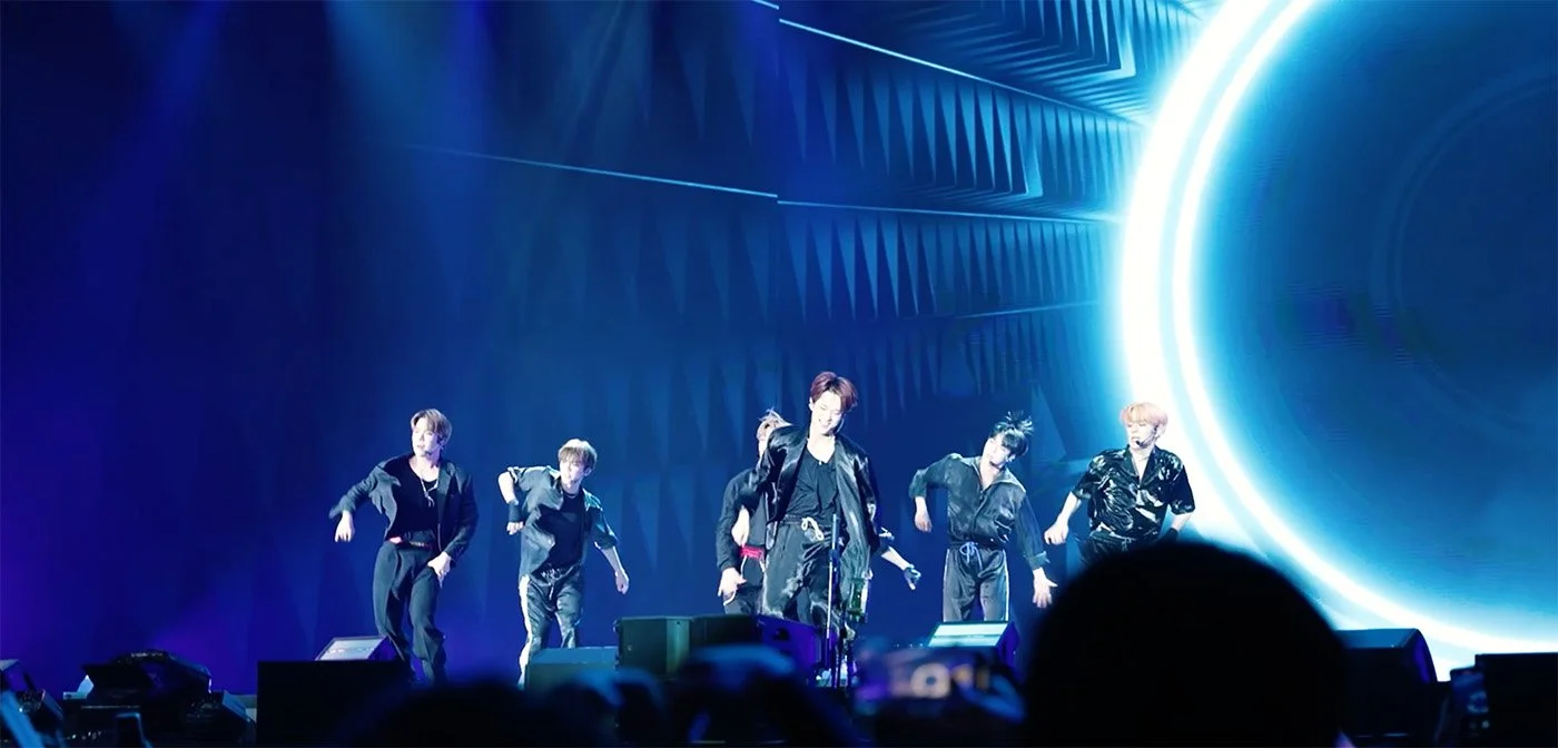 Six performers on stage dancing in front of a large digital screen with a circular bright light and blue abstract patterns.