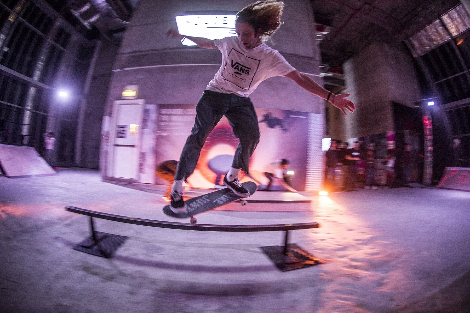 A young male skateboarder with long hair, wearing a white Vans t-shirt and black pants, performing a trick on a rail inside an indoor skate park with colorful lighting and a large screen in the background.
