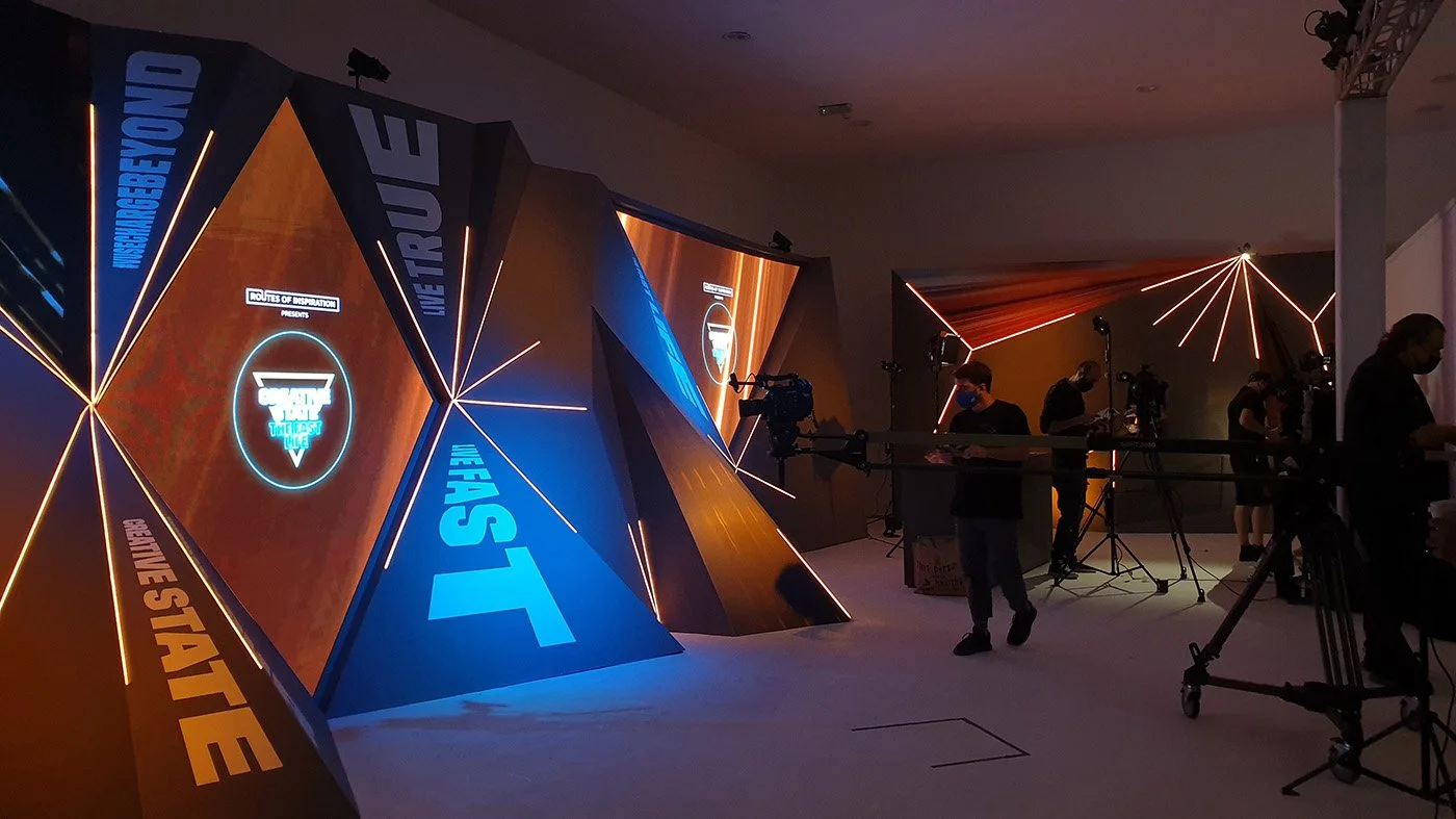 A stage set for a tech or video game event with large geometric screens displaying colorful graphics and text, including 'METRO' and slogans like 'We Are Strong' and 'We Start'. There are multiple people and camera operators preparing equipment in a dimly lit room with neon lighting accents.