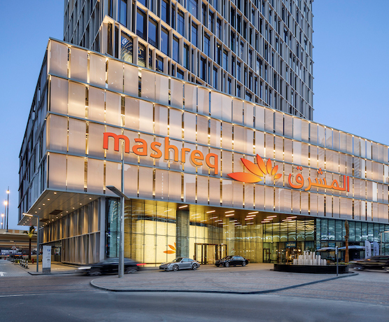 Modern building with illuminated facade and a sign that reads 'Mashreq' in English and Arabic, with a logo of an orange lotus flower, parked cars in front.