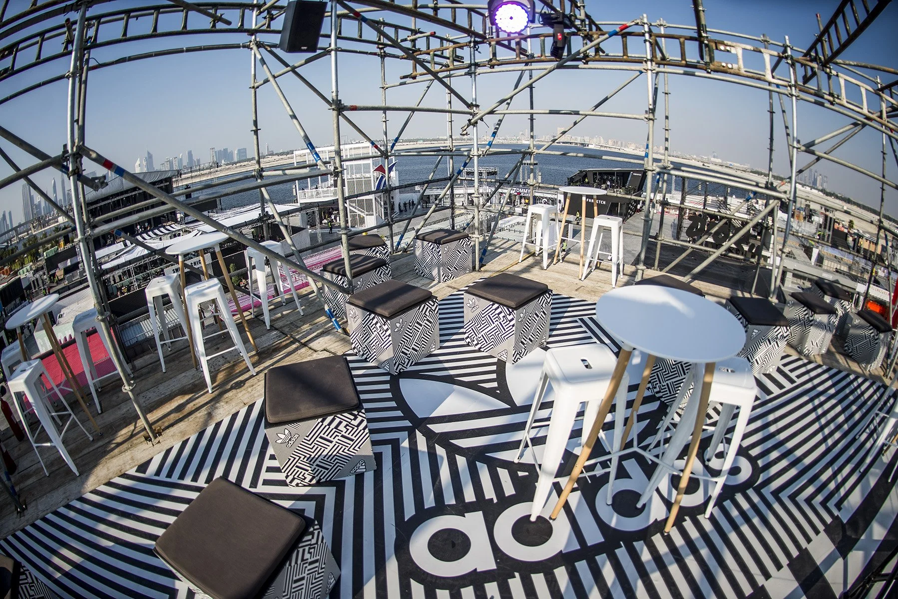 A rooftop bar with black and white patterned furniture and tables, overlooking a city skyline and water. The floor has geometric black and white designs, and there are white bar stools and high tables. Scaffolding structures are present, with a view of a bridge and skyscrapers in the distance.