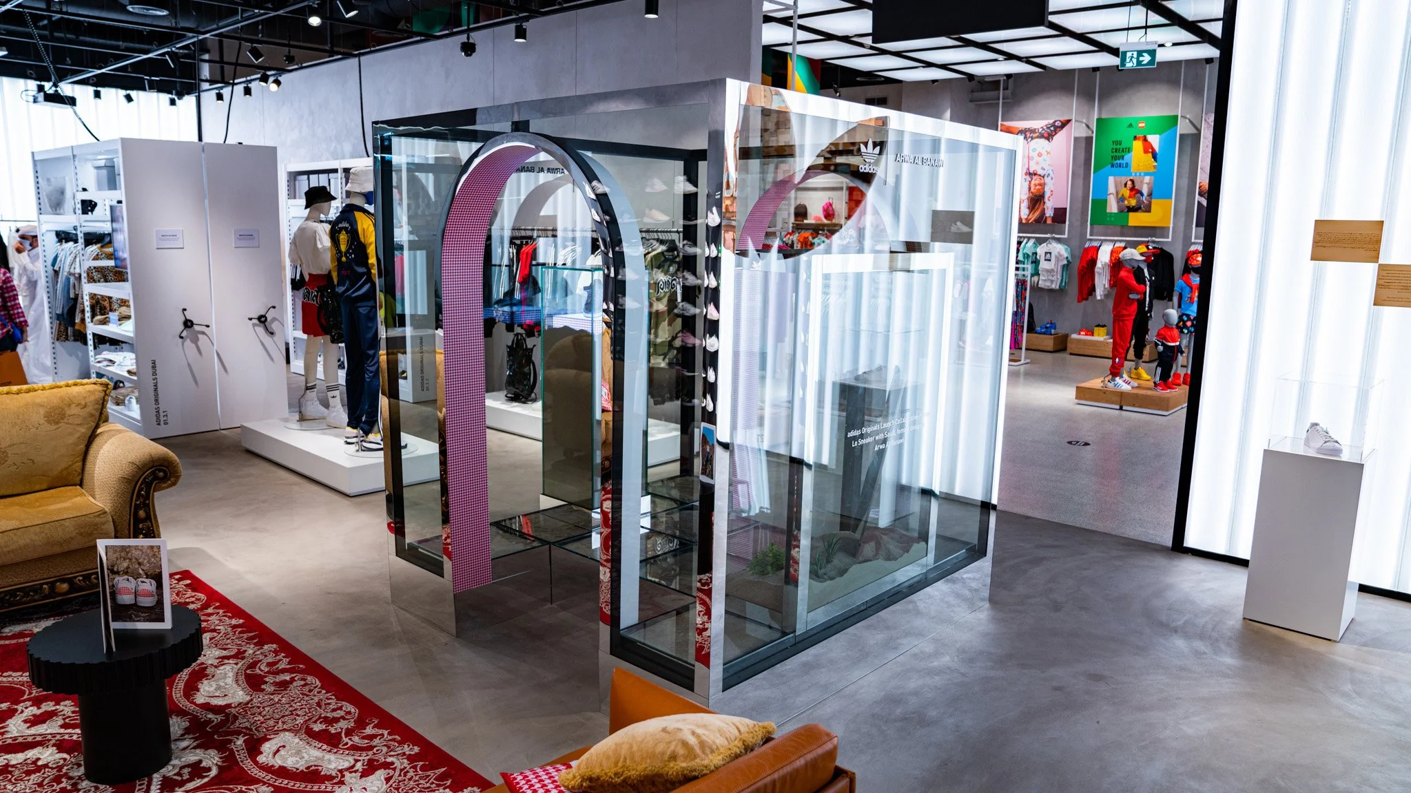Interior of a modern retail store with mannequins dressed in athletic and casual clothing, sneakers displayed in glass cases, and colorful promotional posters on the walls. There is a red patterned rug, a sofa, and a small black table in the foreground.