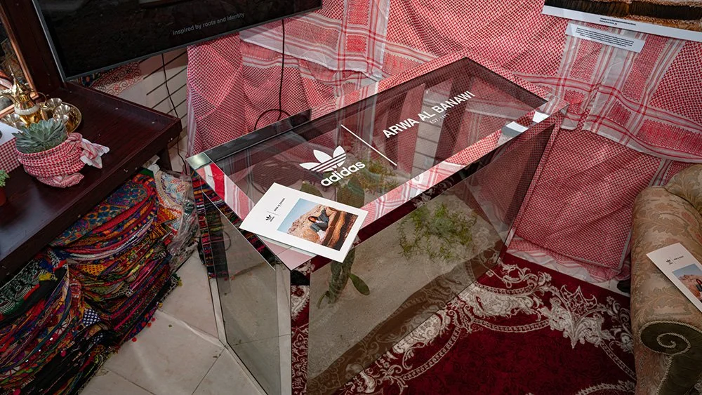 Glass display case with Adidas and Arwa Al Banawi branding, showcasing sneakers, in a decorated room with patterned fabric drapes and colorful textiles.