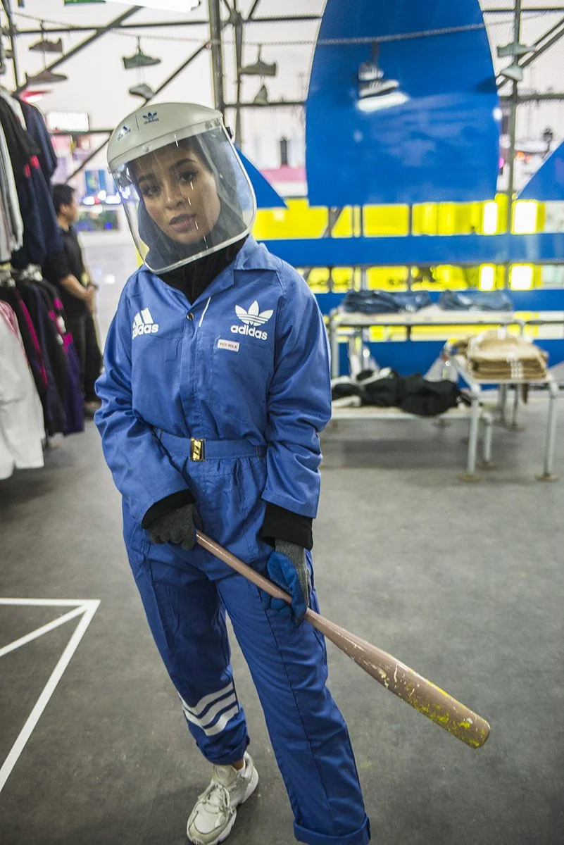 A woman wearing a blue Adidas jumpsuit, a helmet with a face shield, black gloves, and white sneakers stands inside a retail store, holding a baseball bat. The store has clothing items and race track background elements.