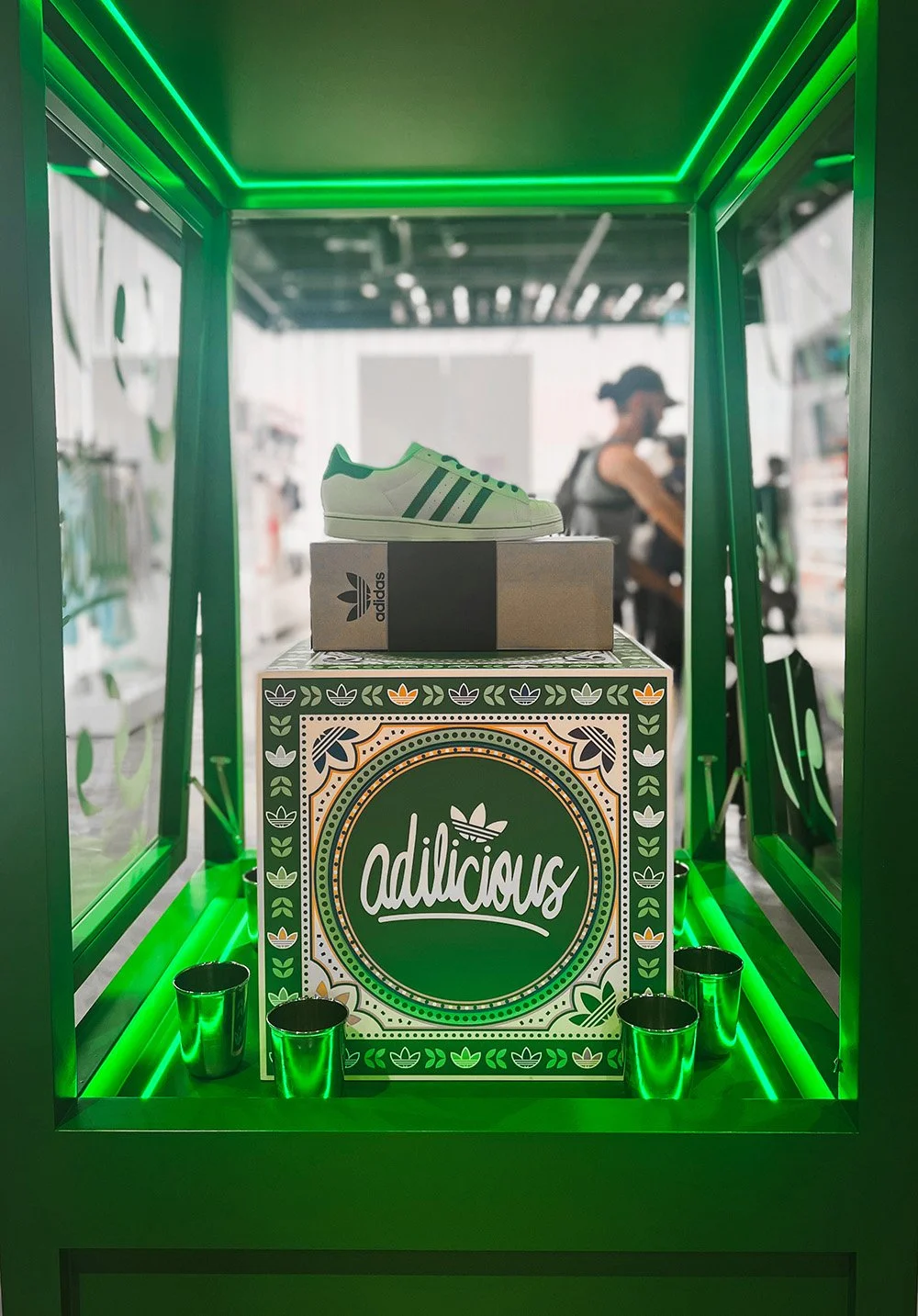 Display of a white Adidas sneaker with green stripes on a shoebox inside a green-lit retail display case, with a woman shopping in the background.
