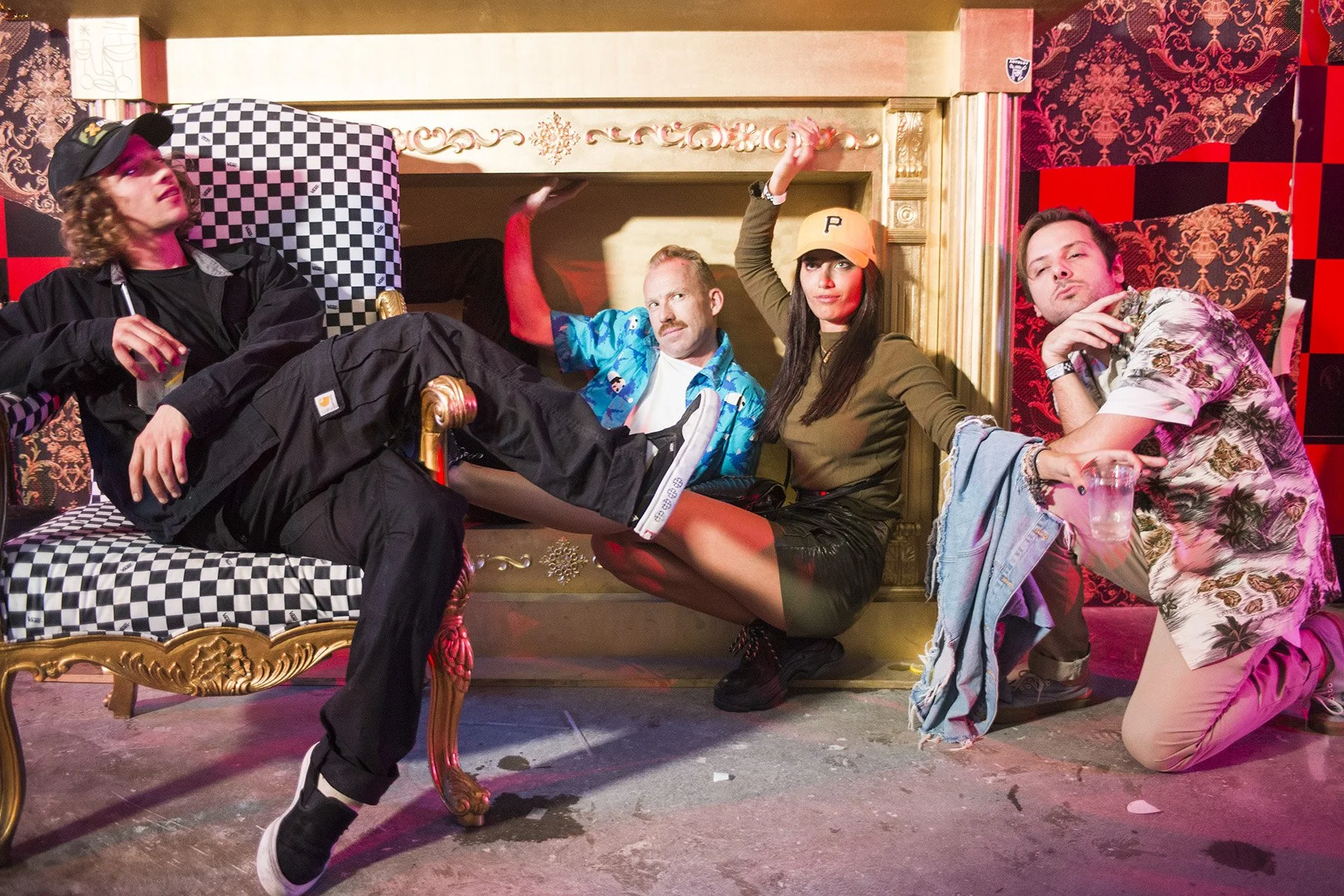 Four young adults sitting and posing on an ornate vintage-style sofa with a checkered and gold design in a decorated room with red and black wallpaper. One person is lying across the sofa, and three others are sitting or kneeling beside them, with relaxed and playful expressions.