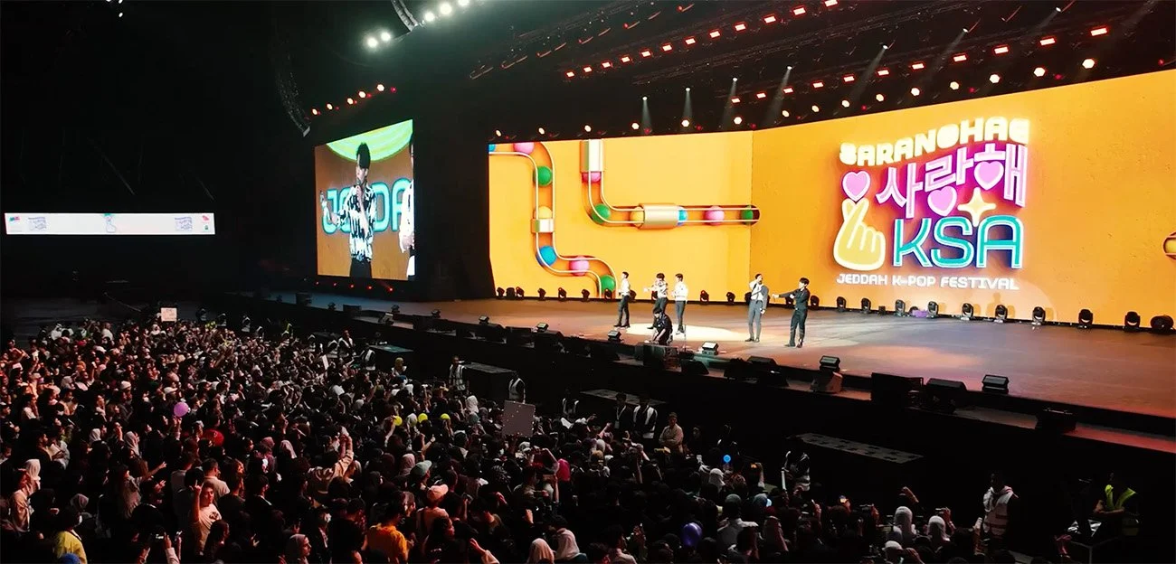 Large stage at a K-pop festival with performers and a massive audience, bright colorful signage in the background, and large screens showing performers.