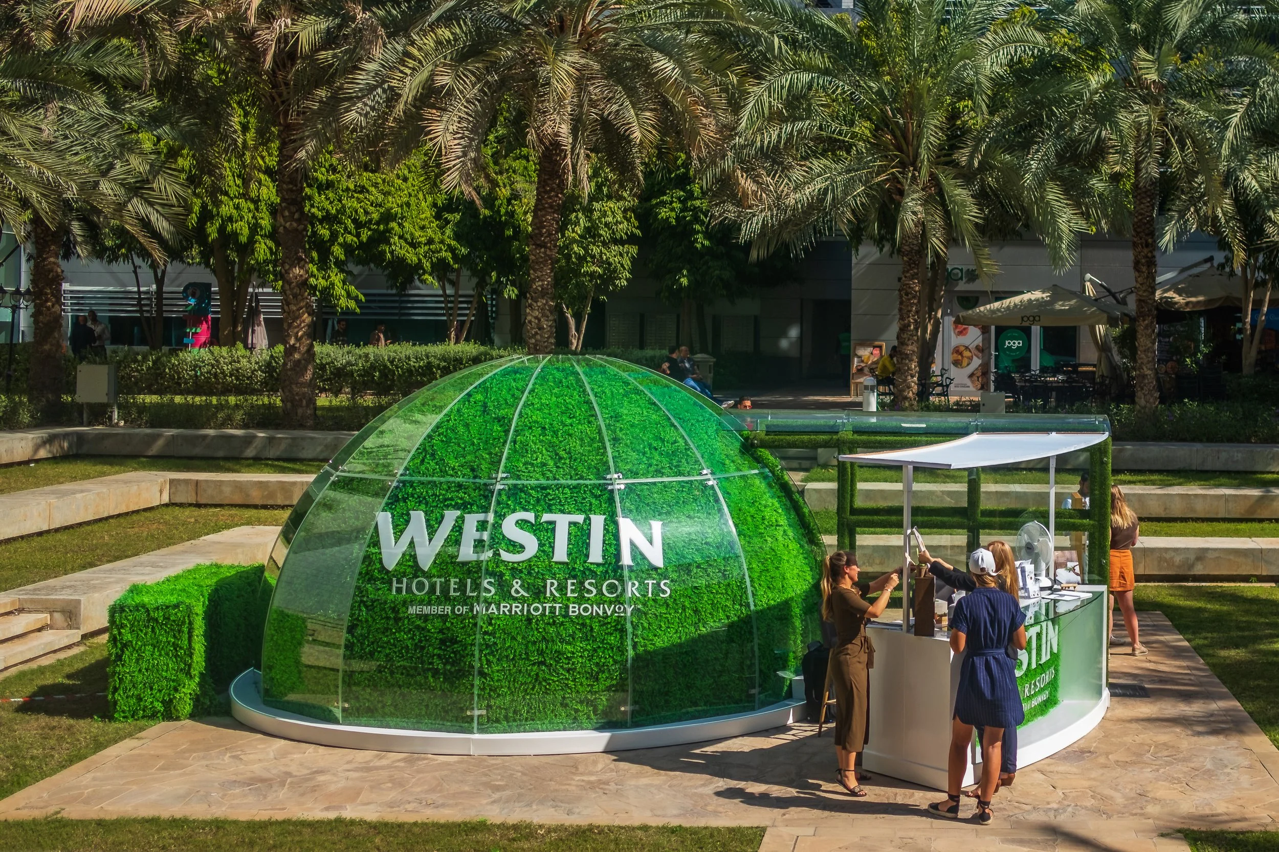 A green, dome-shaped structure with the Westin Hotels & Resorts logo at a public outdoor event. Several people are gathered around the booth, some talking, and others working at the table.