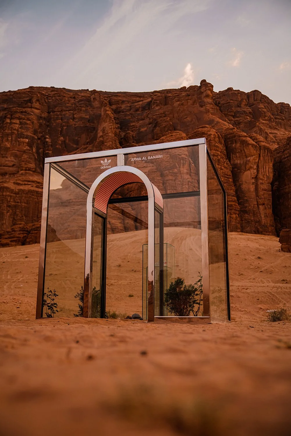 A modern, transparent glass structure with an arched entrance, located in a desert with rocky red cliffs in the background and a clear sky overhead.