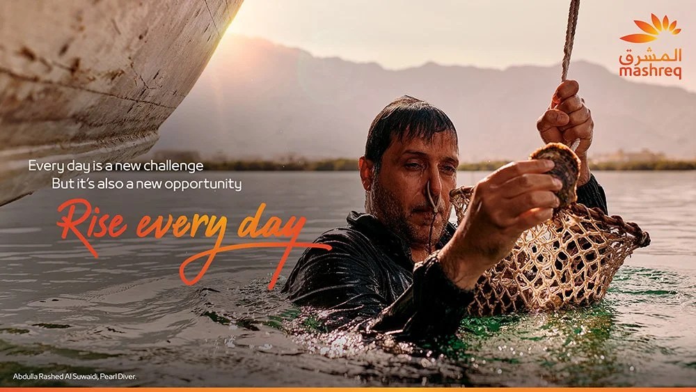 A man in water holding a fishing net with a fish, near a boat at sunset, with mountains in the background. The image includes text saying 'Every day is a new challenge but it's also a new opportunity' and 'Rise every day.' The Mashreq logo is in the top right corner.
