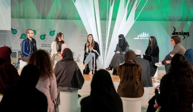 Panel discussion with six women on stage, four women with covered faces, and a man in a blue jacket, at an event with audience members seated and listening.