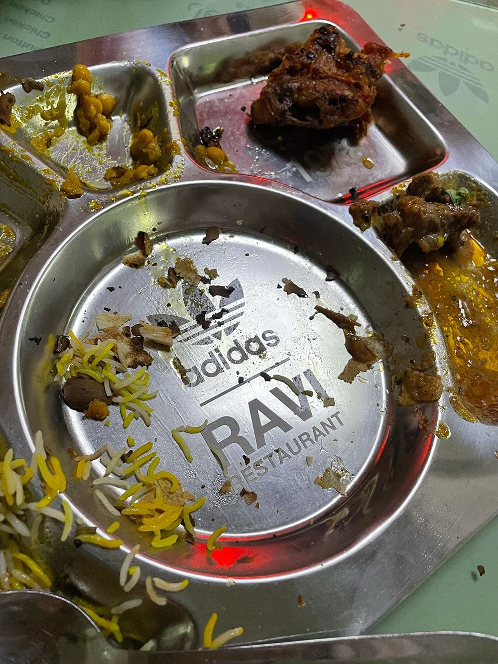 Empty stainless steel tray with remnants of food, including rice and vegetables, inside a restaurant named 'TRAVE' with an Adidas logo.