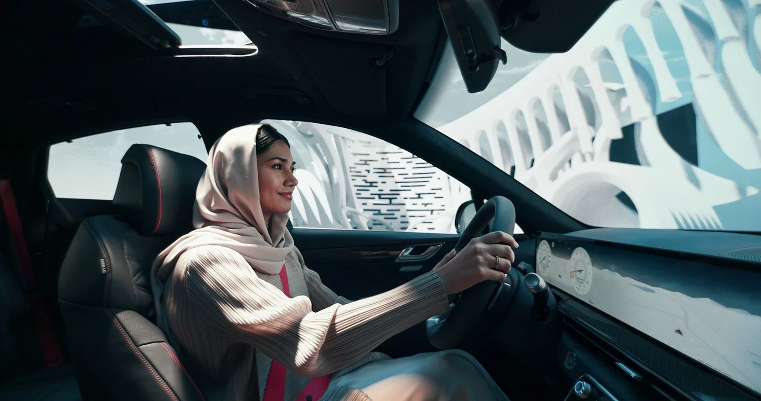 A woman with a beige headscarf driving a car, smiling, with a futuristic white structure visible through the windshield.