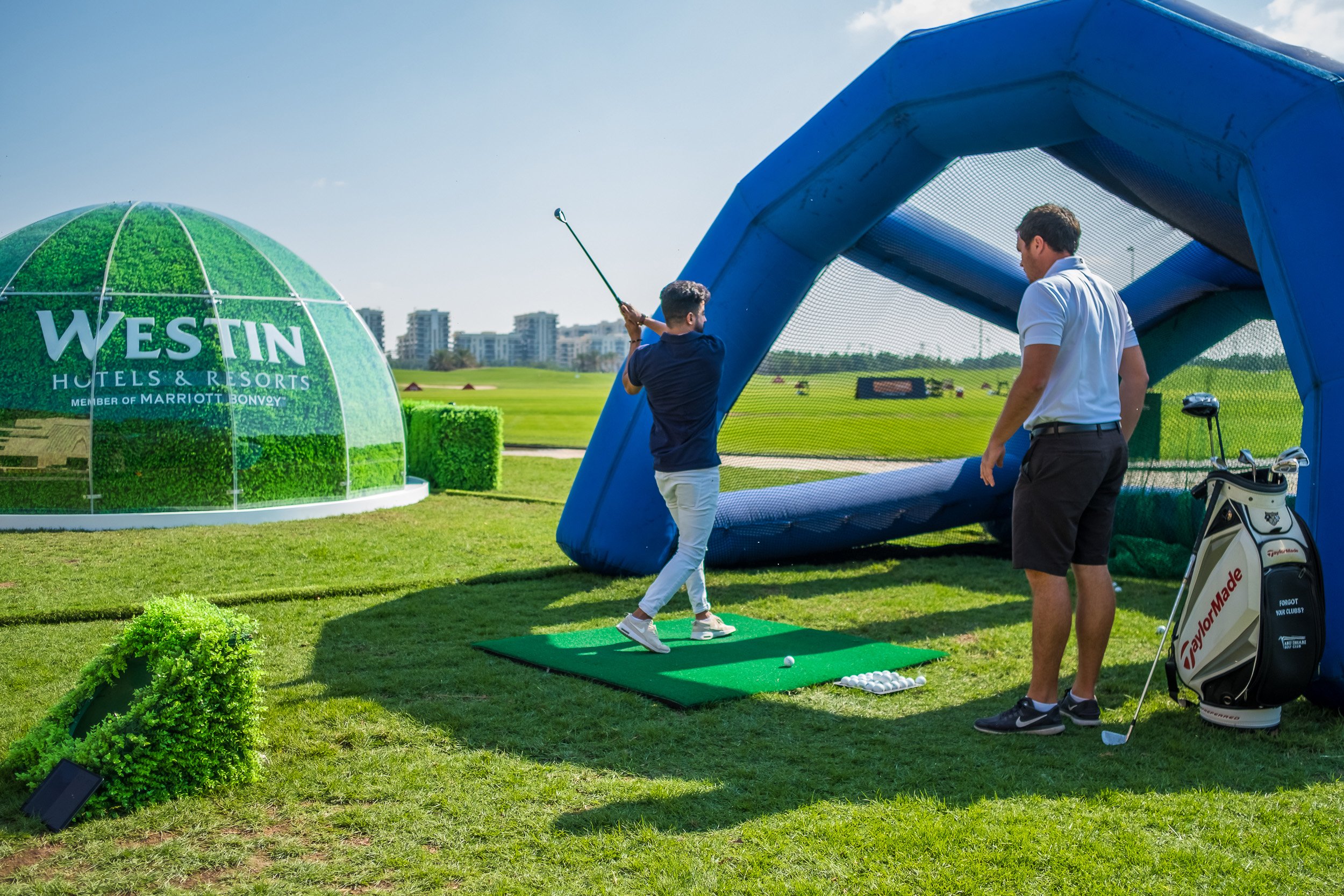 Two men practicing golf on a driving range under a blue tent with a golf ball on a tee. A large green inflatable dome with the text 'Westin Hotels & Resorts' is visible in the background.