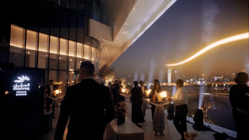 An outdoor rooftop event at night with people socializing, modern decor, city skyline, and water view, illuminated by lights.