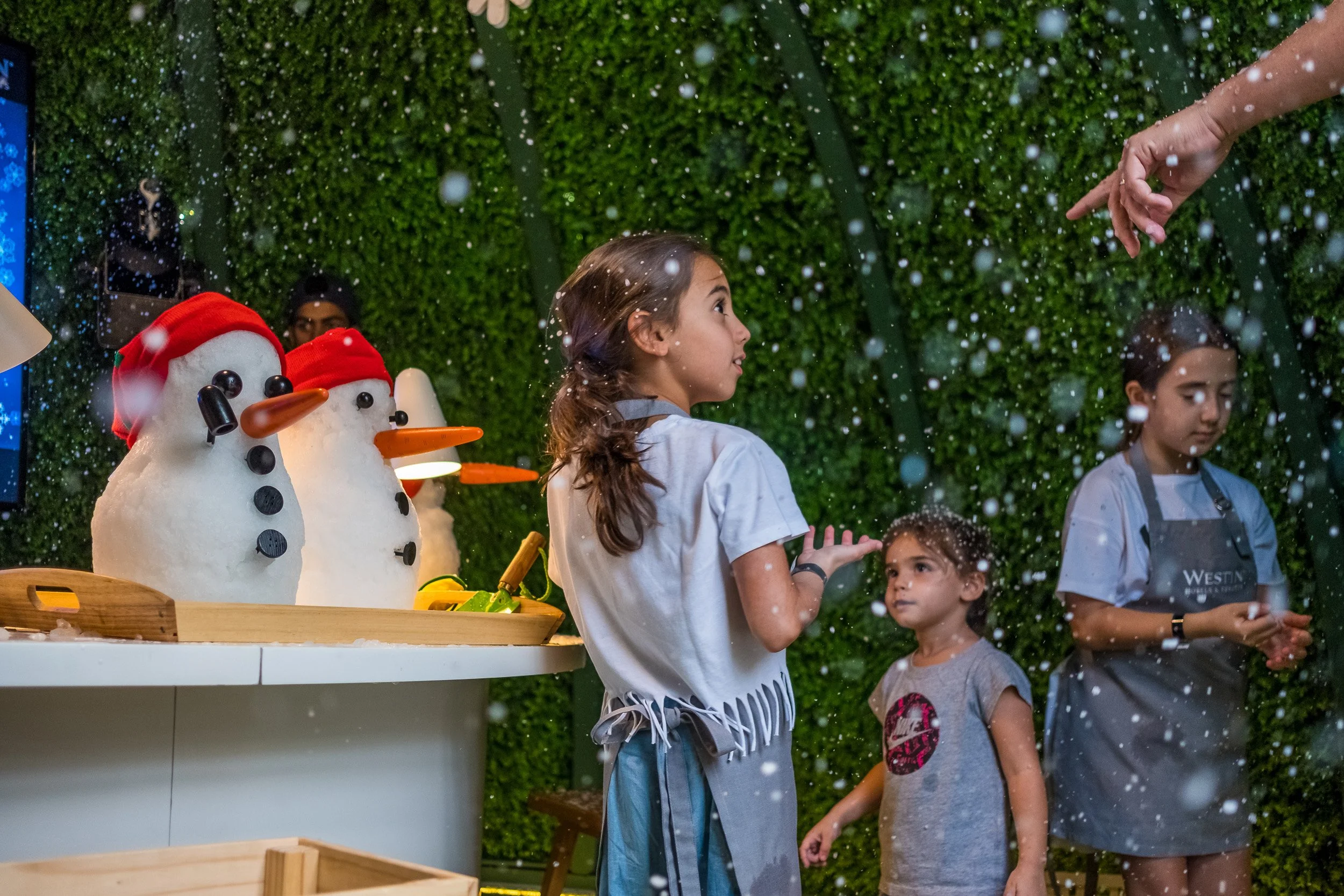 Children participating in a winter holiday event with snowmen decorations and artificial snow, standing in front of a green leafy wall.