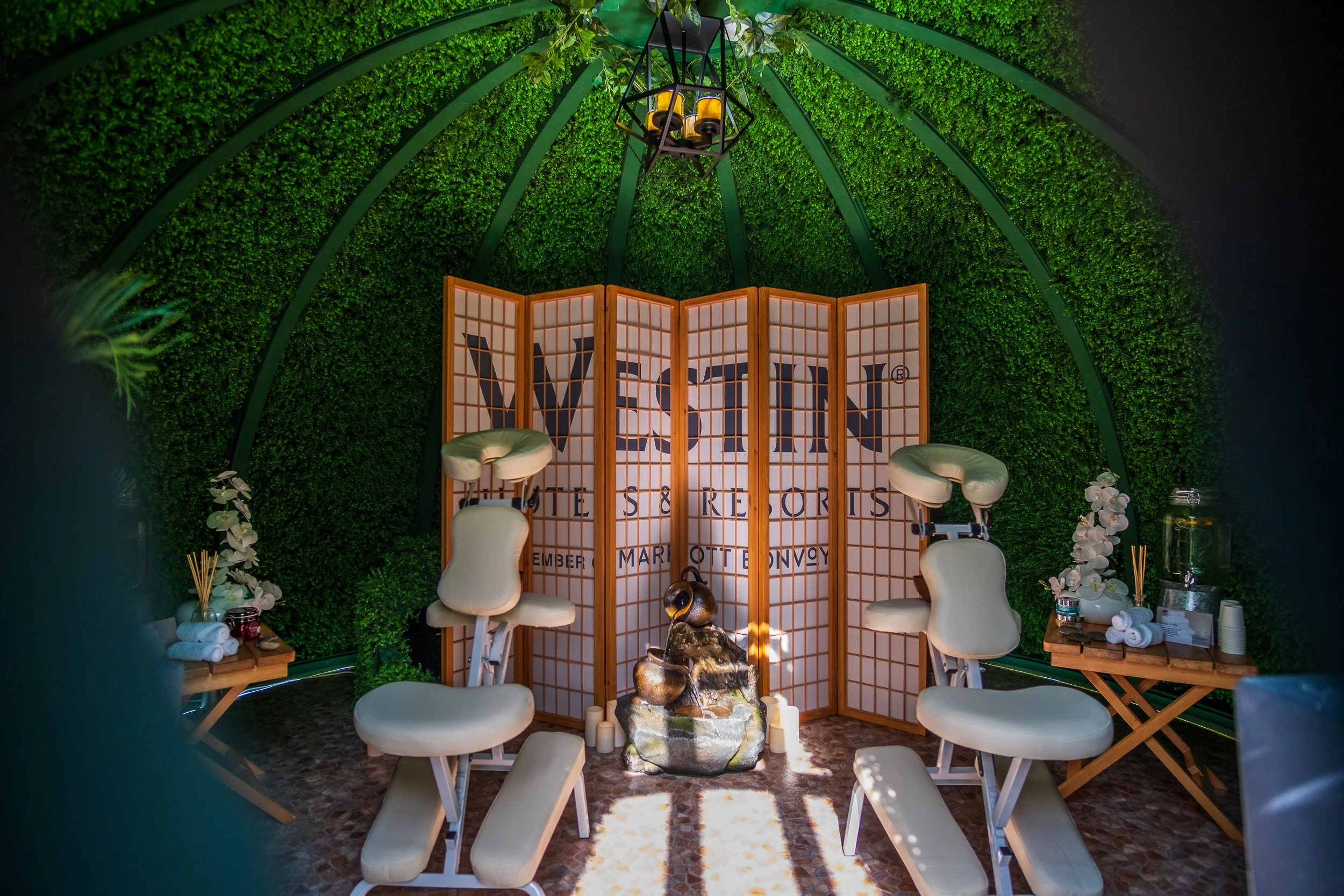 Indoor event space with green foliage walls, a folding screen with the words 'VEX' and 'Venture Sports' partially visible, seating with white chairs, floral arrangements, candles, and decorative objects, and a modern chandelier hanging from the ceiling.