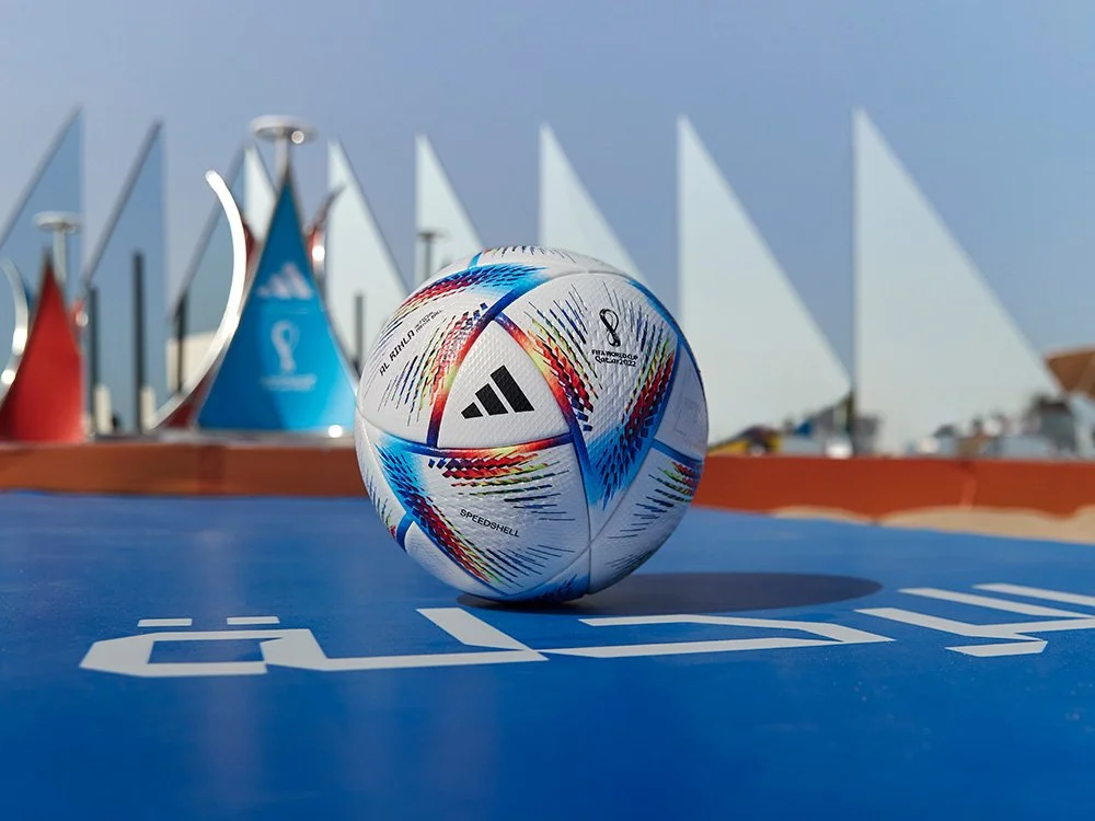 A soccer ball with colorful patterns is on a blue surface outdoors, with sailboats and a cloudy sky in the background.