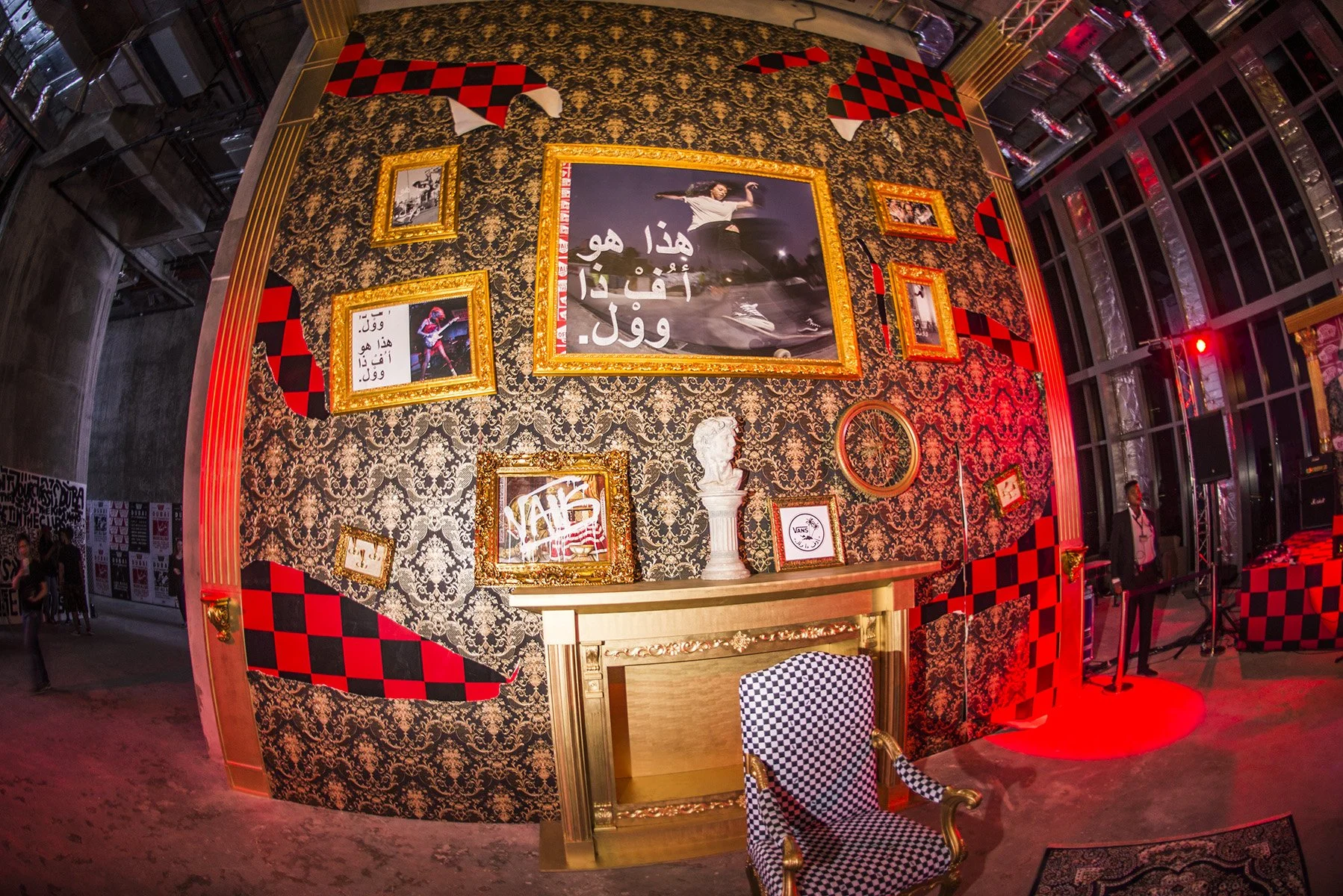 An interior decorated wall with ornate gold frames, patterned wallpaper with black and gold floral design, and red and black checkered accents. Various framed pictures, a bust statue, and a sign reading 'NAIS' are displayed. There is a fireplace with an elaborate gold frame, a checkered armchair, and a man in business attire standing nearby. The setting appears to be at an event or exhibition with red lighting effects.