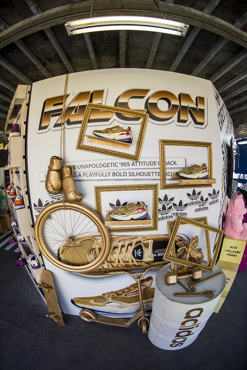 Display of gold-colored Adidas sneakers, boxing gloves, a skateboard, a bicycle, and empty picture frames on a white background with bold black and gold text that says 'FALCON' and Adidas logos.