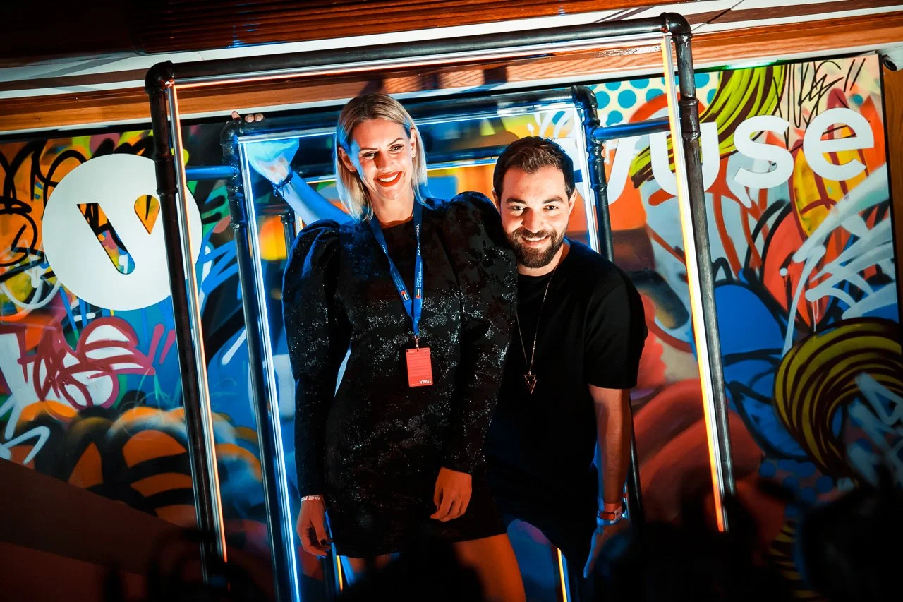 Two people, a woman and a man, smiling and posing together inside a colorful artsy photo booth or exhibit with graffiti-style murals in the background.