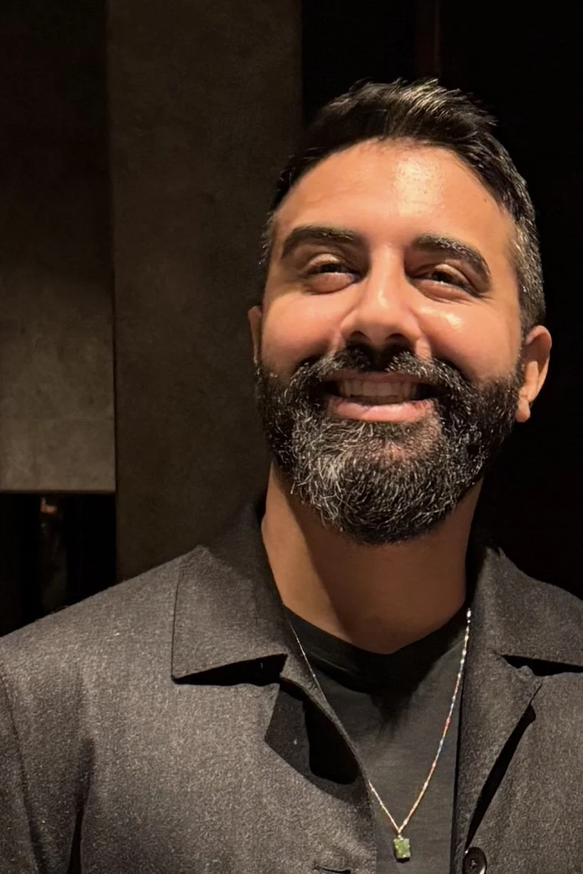 A smiling man with dark hair and a beard, wearing a dark blazer and a necklace with a green pendant, standing against a dark background.