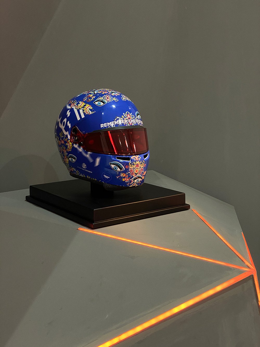 A blue motorcycle helmet with colorful eye patterns and floral designs, displayed on a black stand with orange LED lighting accents.