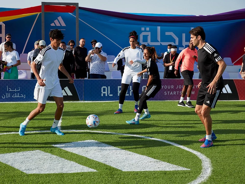 Group of athletes on a football field playing a casual game, with audience members watching in the background.