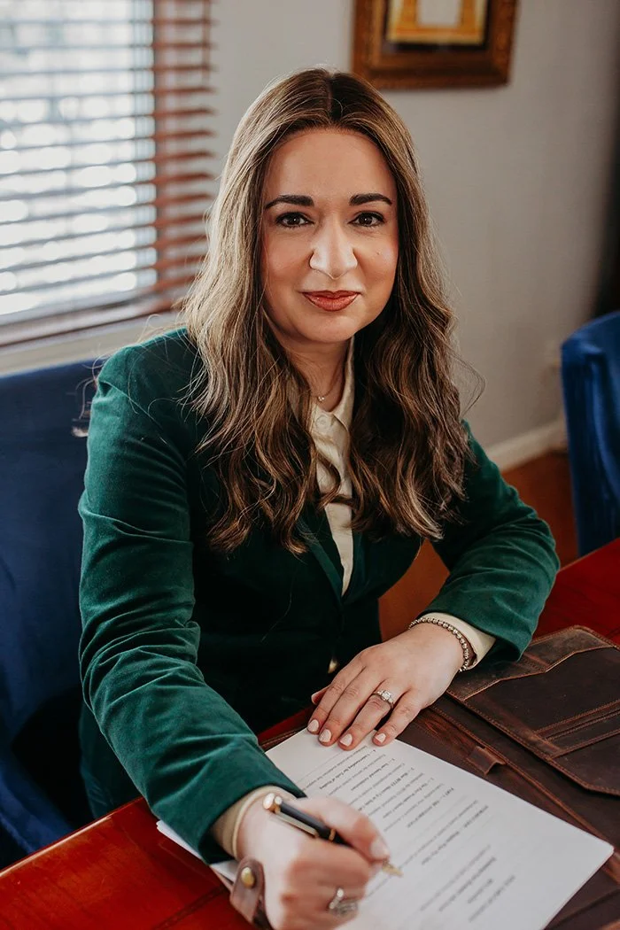 A woman with long wavy brown hair, wearing a green blazer and white shirt, sitting at a desk signing a document, with a window with blinds behind her.