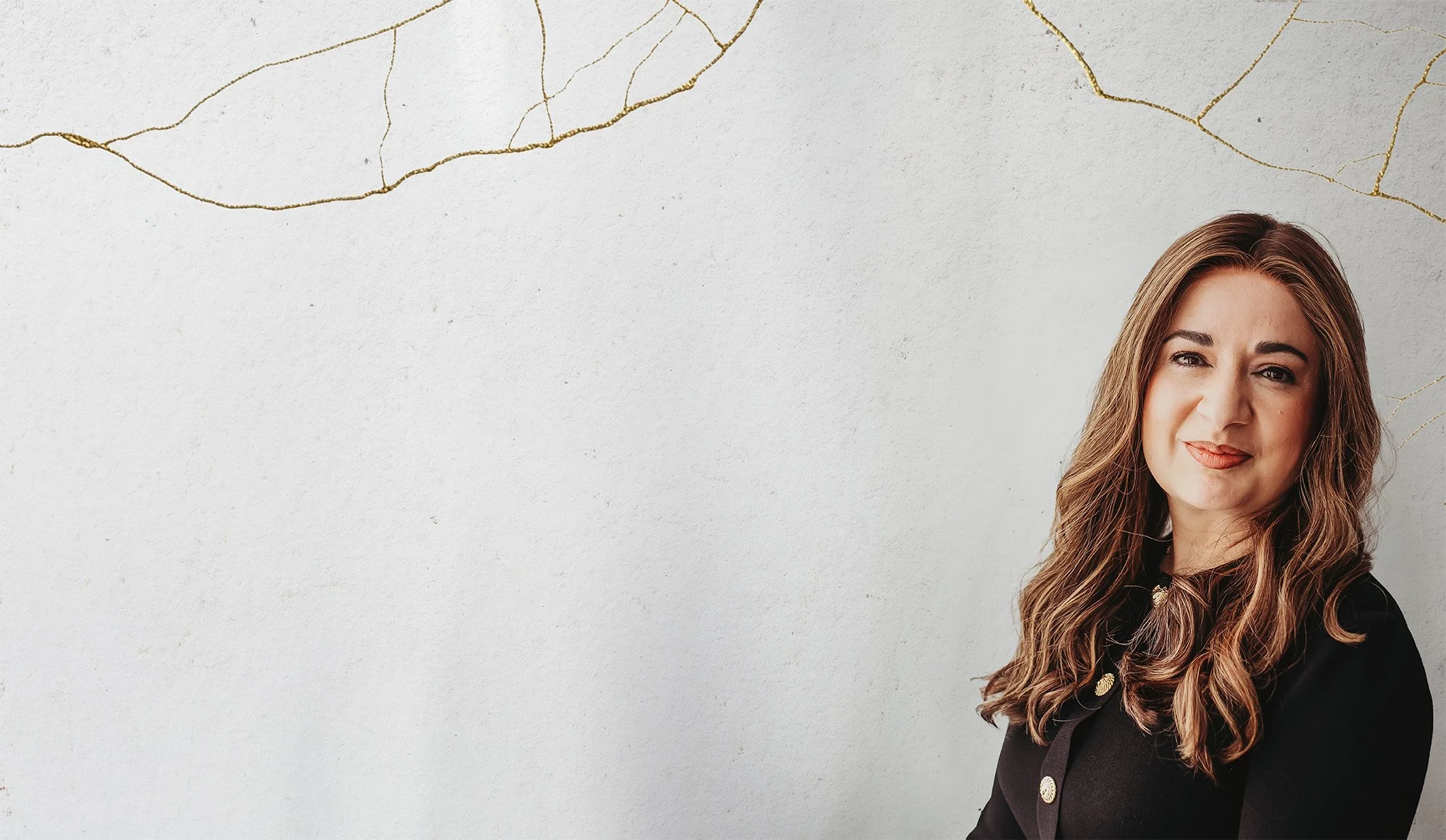 A woman with long wavy hair wearing a black blazer, standing against a white wall with gold cracks.