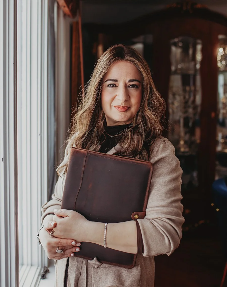 A woman with wavy brown hair holding a dark brown leather folder, standing near a window in a warmly lit room.