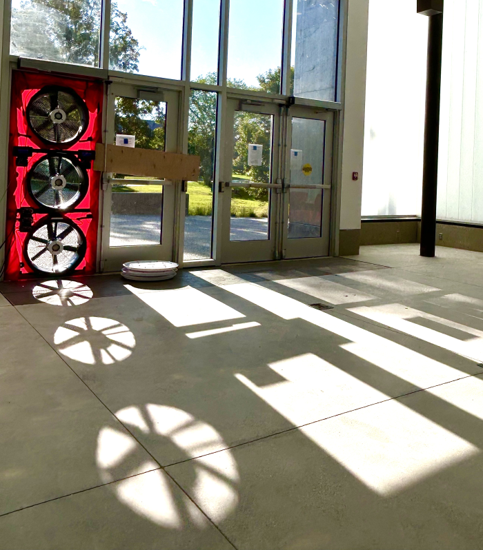 Sunlight streaming through glass doors creating shadow patterns resembling fans on the floor. A red display with four large black fans and a robot vacuum cleaner are inside the building.