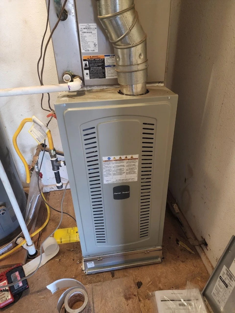 A furnace and HVAC system in a utility room with various pipes, wires, and tools around it.
