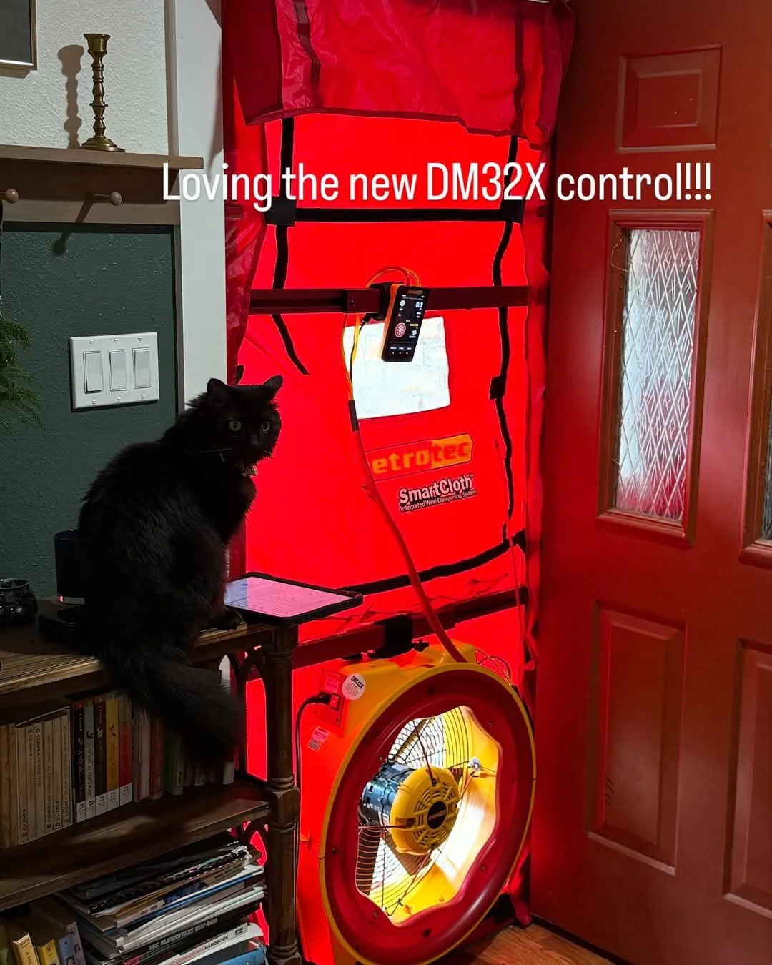A black cat sitting on a small wooden bookshelf next to a large, yellow dryer vent cleaning fan and an orange portable heater. A red SmartDry tarp with a control device hanging in front is set up at the entryway, indicating a new DM32X drying control system. Text overlay says, 'Loving the new DM32X control!!!'