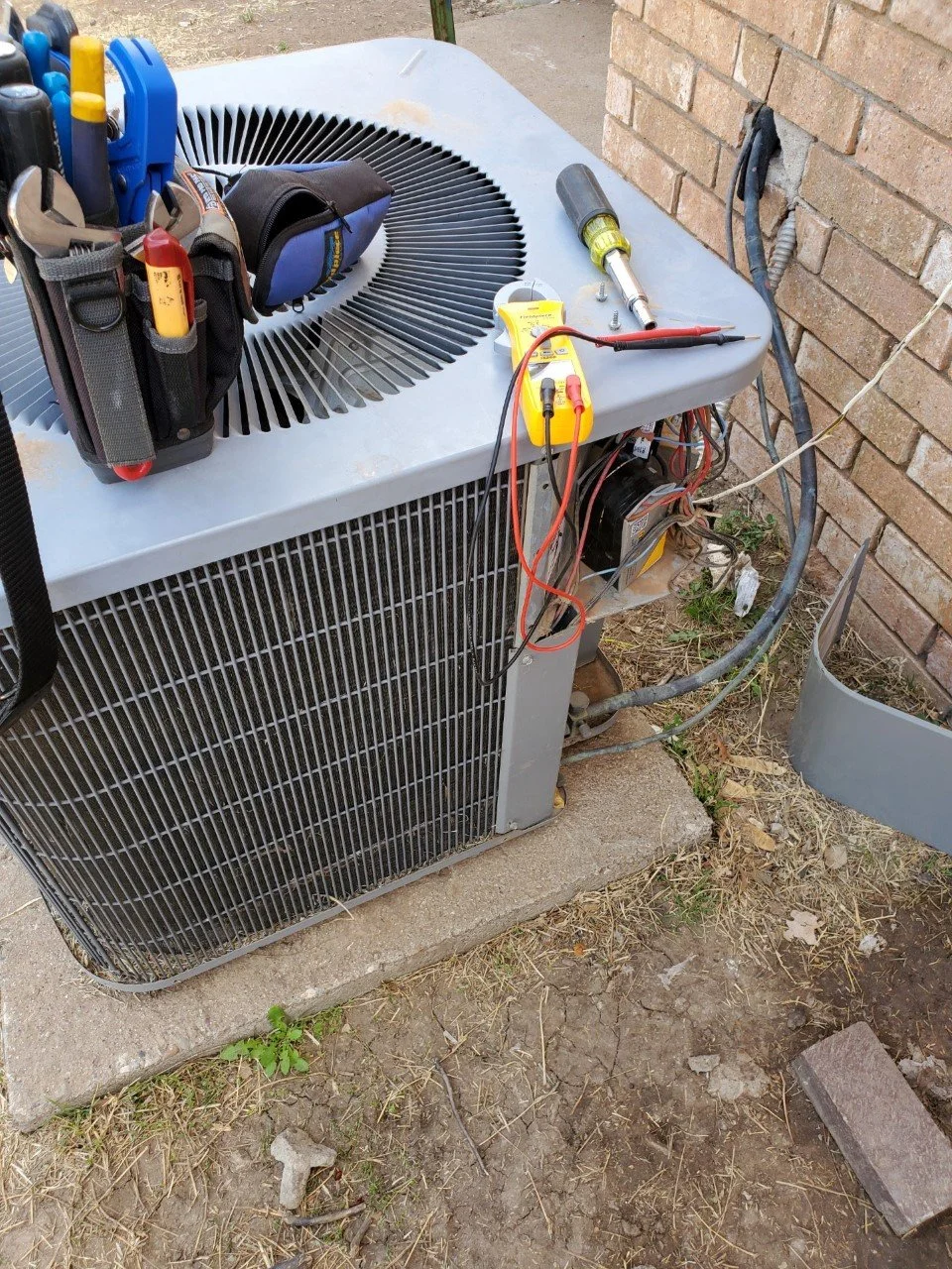 An outdoor HVAC unit with tools and a multimeter placed on top, located beside a brick wall and on a concrete slab, with electrical wires connected and some exposed ground around.